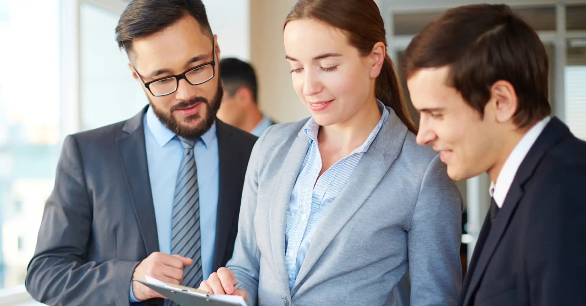 Three business professionals in suits are looking at a tablet screen together in an office setting.