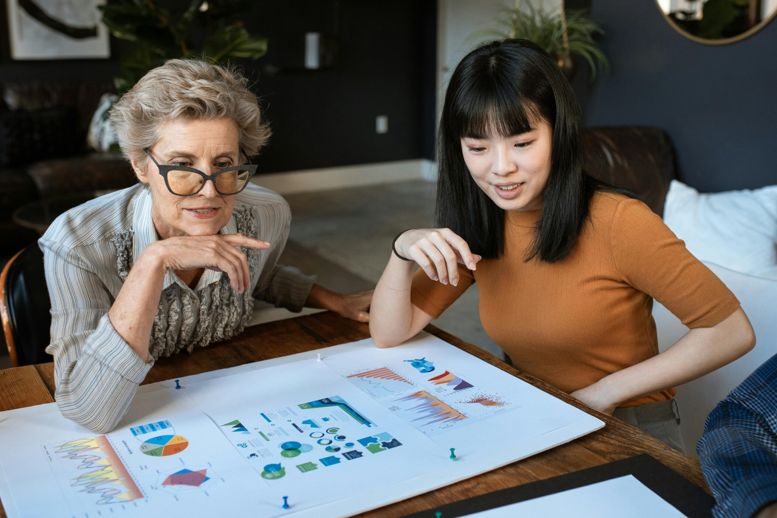 Two women are seated at a table, discussing charts and graphs displayed on large sheets of paper. They appear focused and engaged in conversation, exploring an exciting franchise opportunity.